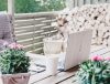 Laptop and coffee cup on a wooden patio table with plants and a stack of firewood in the background