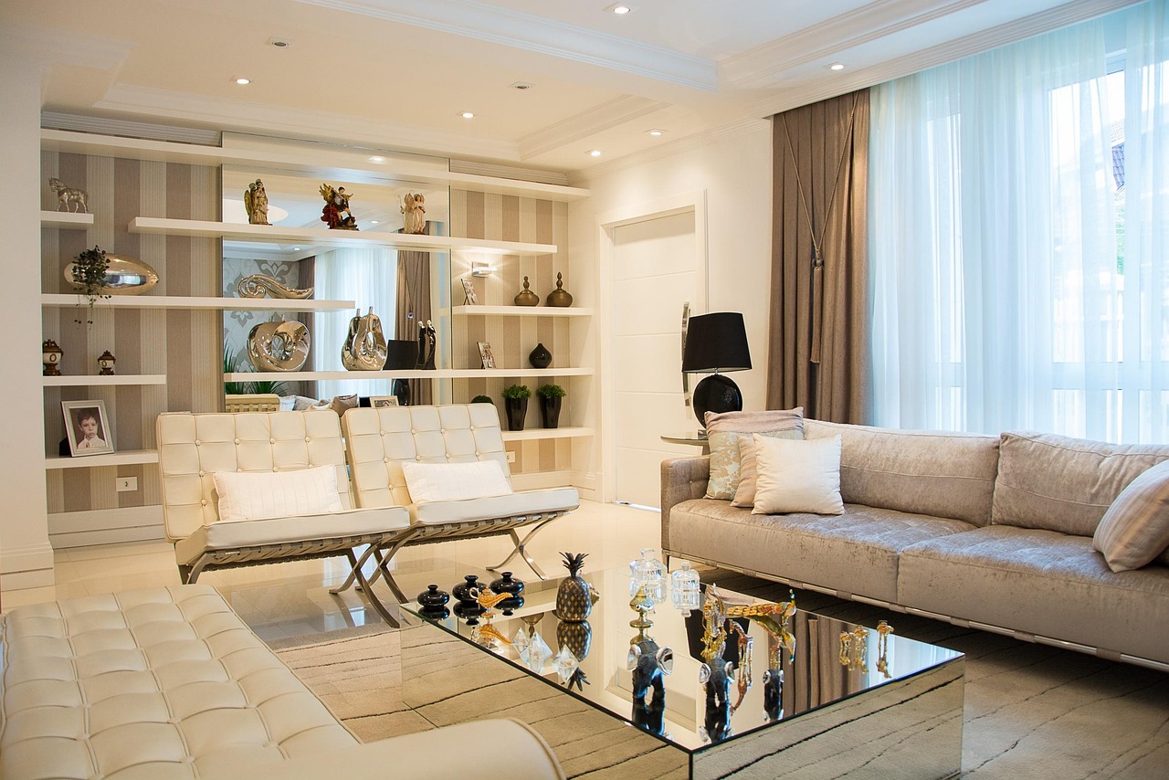 bright, luxurious living room with cream and beige tones featuring a plush velvet sofa, two modern white chairs, and a mirrored glass coffee table decorated with crystal ornaments. Built-in wall shelving displays elegant vases, sculptures, and family photos