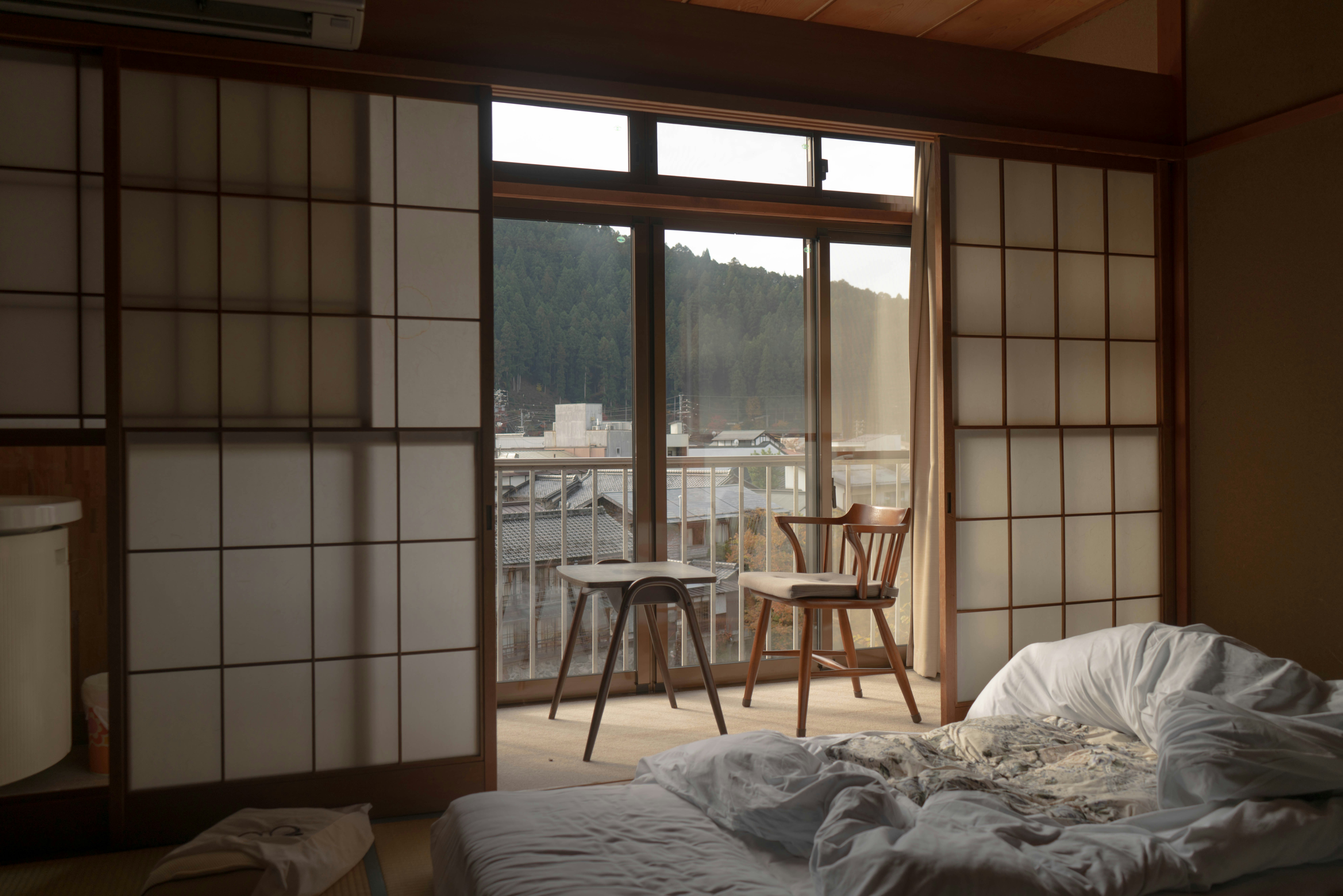 serene Japanese-style room with sliding shoji doors, a low unmade futon bed on a tatami mat floor, and a view of a forested hillside through large windows. Two wooden chairs and a small table sit on the balcony, overlooking traditional rooftops and a quiet mountain town