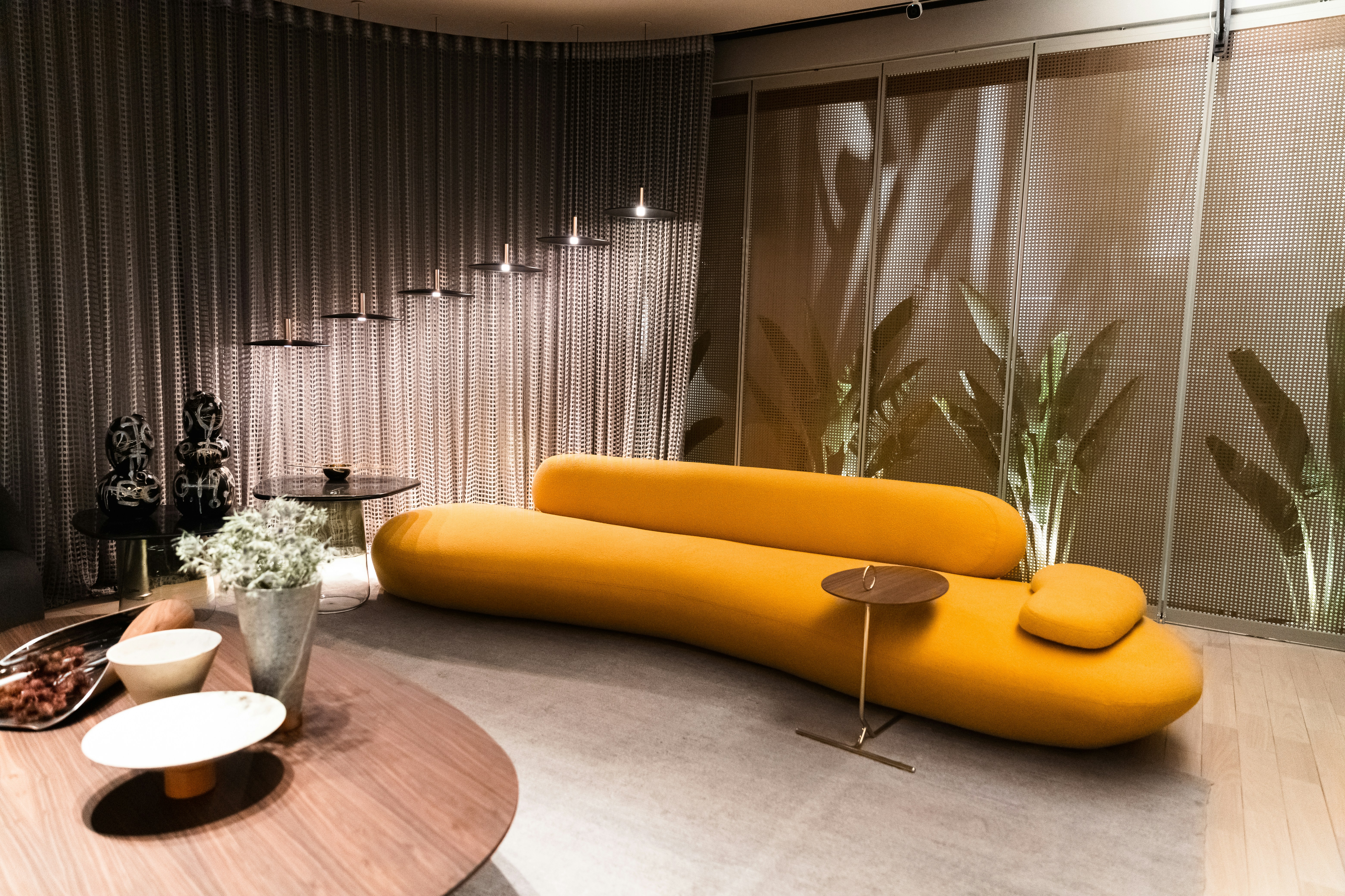 modern, minimalist lounge with a striking mustard-yellow curved sofa, soft grey rug, and round wooden coffee tables. The room features artistic lighting, potted greenery, and a perforated metallic wall with soft shadows from tall plants behind.