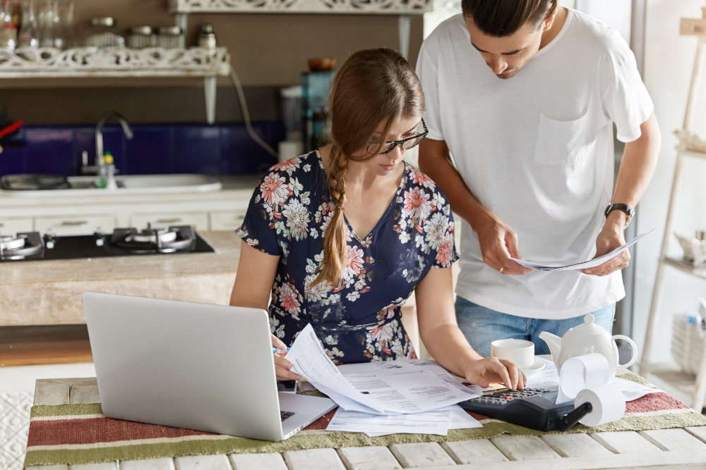 A man and woman collaborate on paperwork at a kitchen table, surrounded by various kitchen items